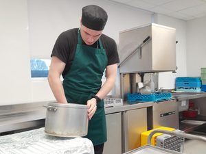 Riley in the kitchen at Mayfield school, wearing dark green apron with hand in a large cooking pot
