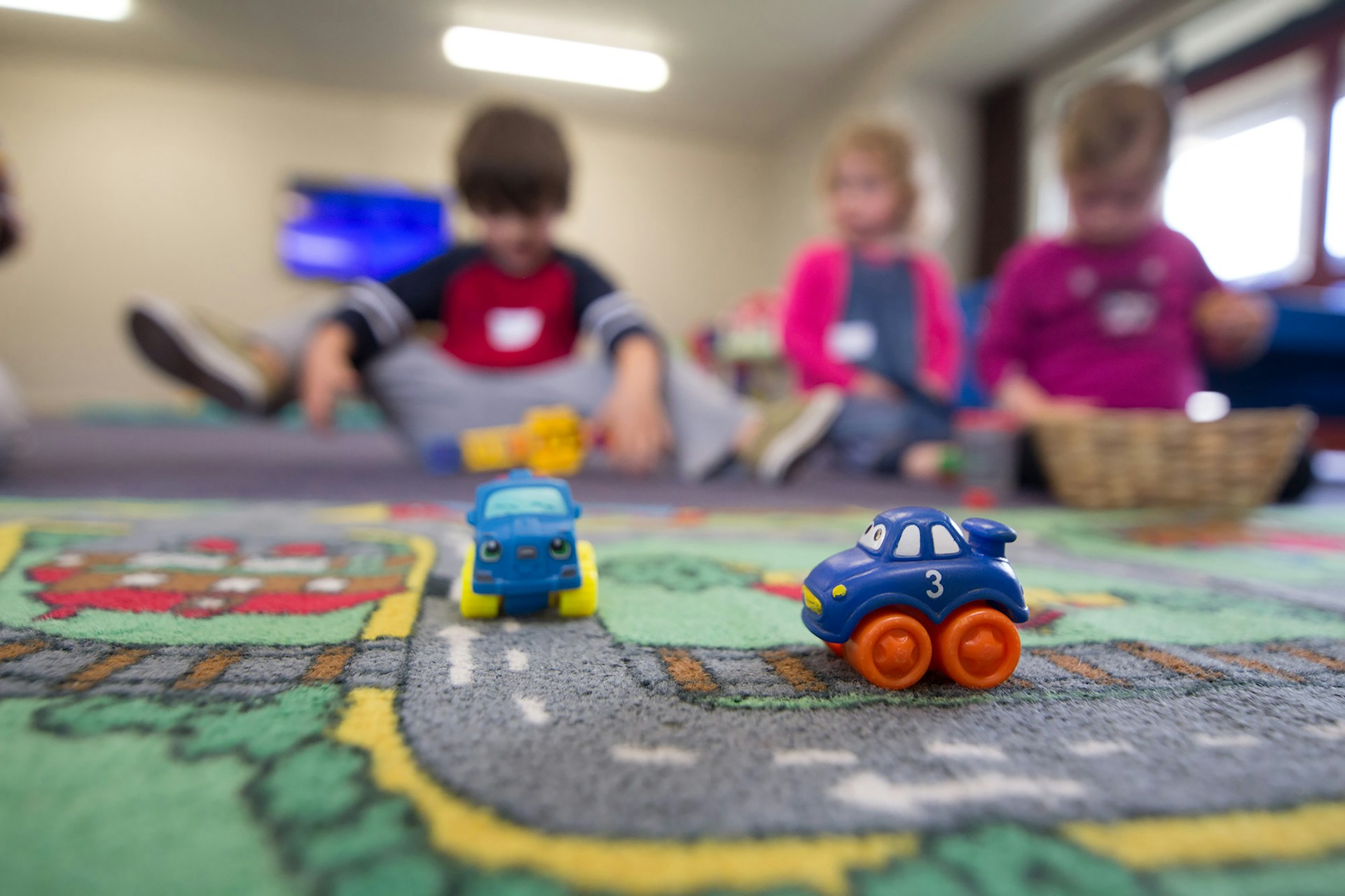 Toy cars in foreground with blurred children in nursery sitting on floor with sensory baskets. 
