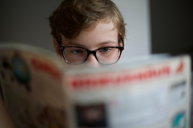 Boy with glasses reading newspaper in front of his face