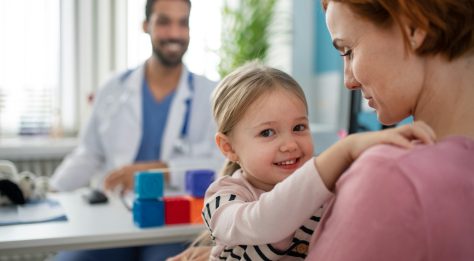 smiling small child looking over parent's shoulder, with health professional in the background