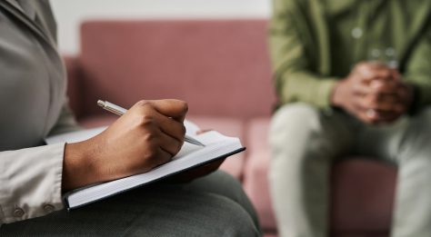 close up of person writing in notebook, with blurred view of someone sitting on a sofa behind